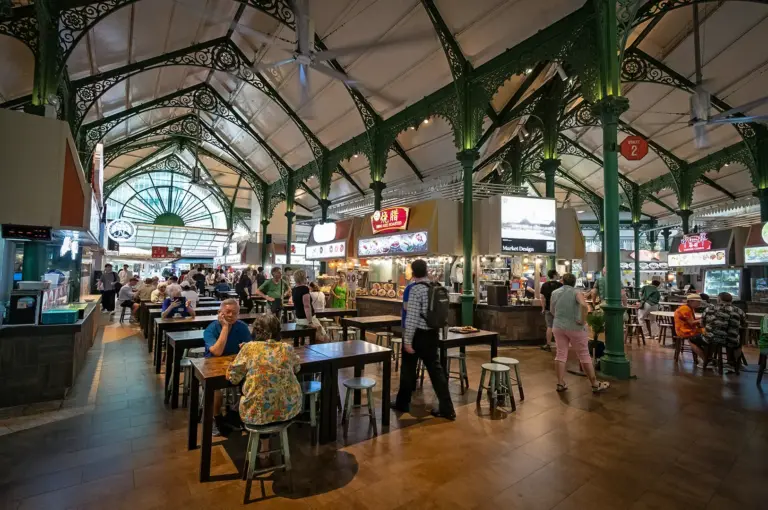 Spacious Singapore hawker centre with heritage architecture, bustling food stalls, communal dining tables, and visitors enjoying a wide variety of local street food.