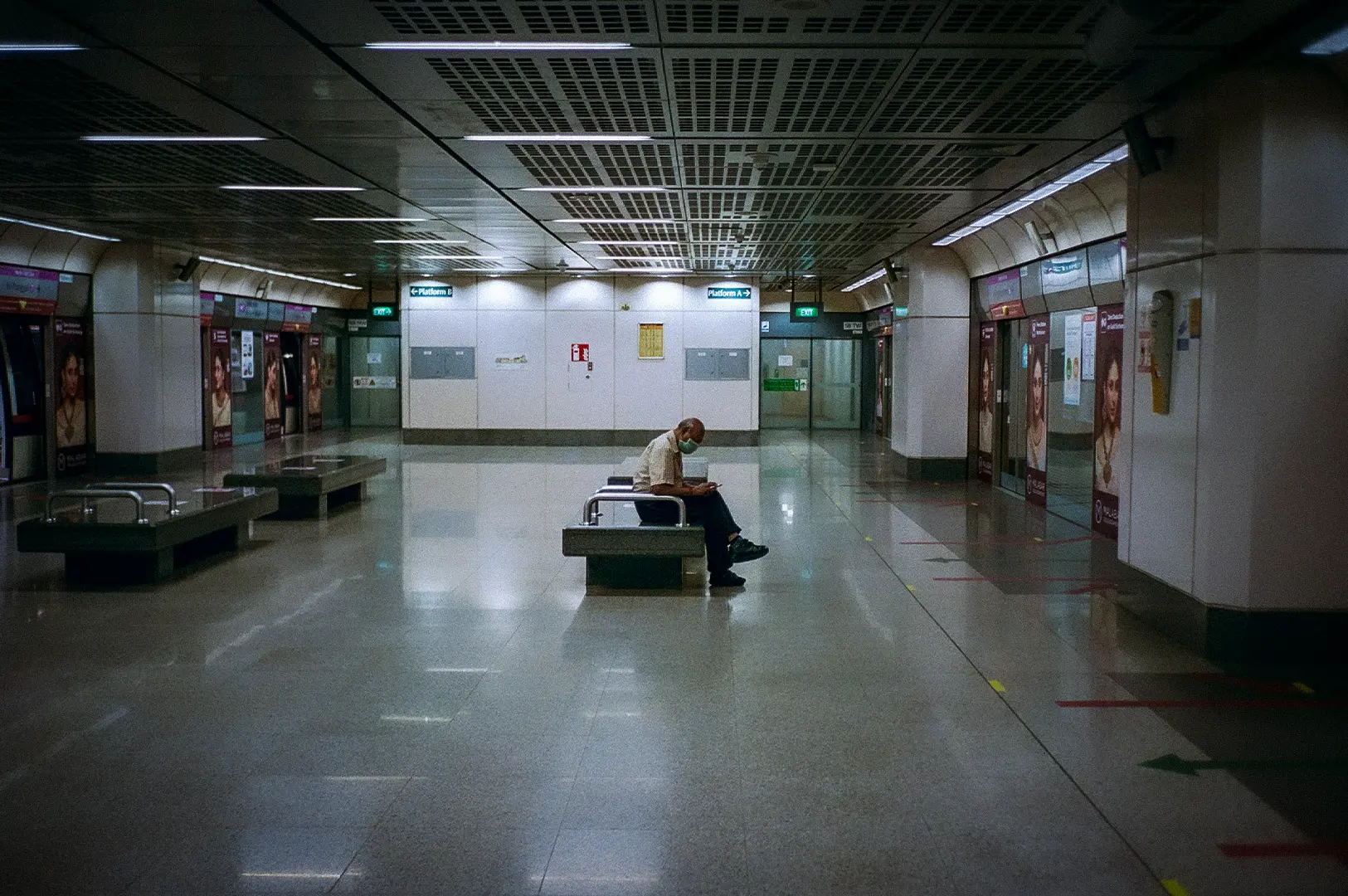 Interior of a Singapore MRT station platform featuring spacious waiting areas, platform screen doors, overhead directional signage, and a commuter seated on a bench.