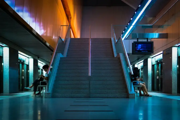 Wide-angle view of the staircase inside Dhoby Ghaut MRT Station in Singapore, featuring modern architecture, platform entrances, and commuters waiting underground.