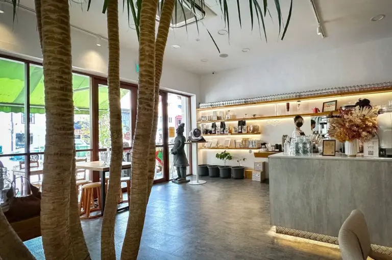 Modern interior of Kings Cart Coffee Factory in Singapore featuring a minimalist coffee bar, warm lighting, indoor trees, and spacious seating for a relaxing café experience.