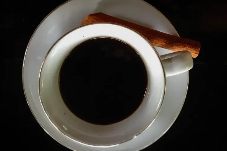 Traditional Singapore kopi served black in a white cup with a cinnamon stick on the saucer, photographed from above on a dark wooden table.