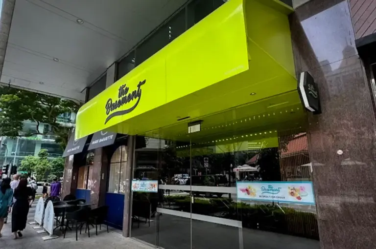 Modern exterior of The Basement Cafe in Singapore featuring its distinctive lime green storefront, glass entrance, and outdoor seating along a busy city street.