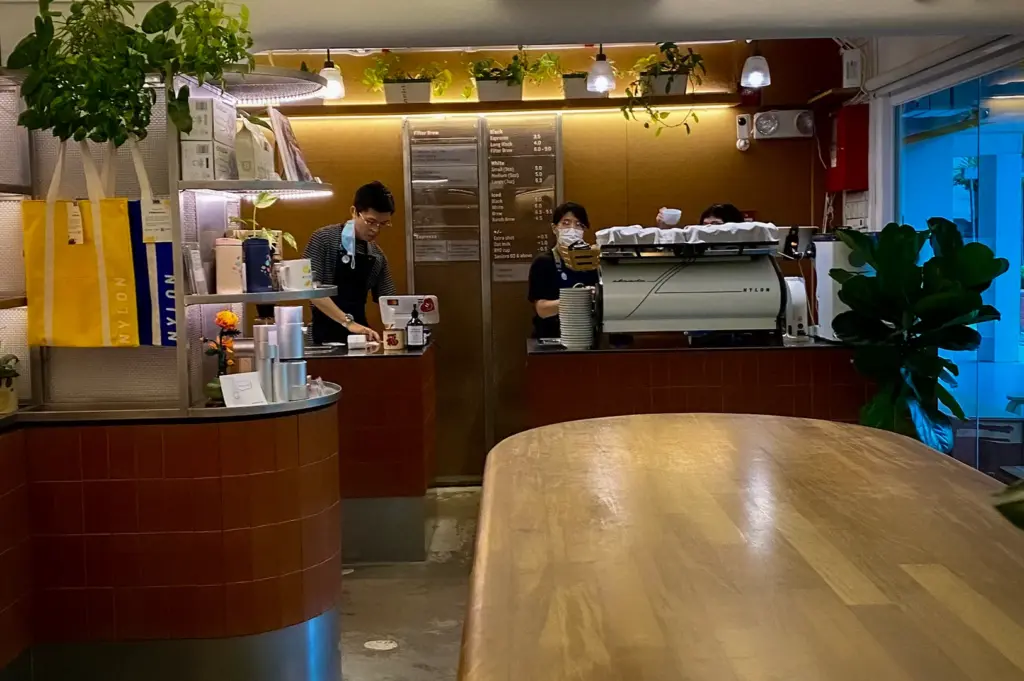 Warm and modern interior of Nylon Coffee Roasters Singapore with baristas preparing specialty coffee, wooden communal table, espresso bar, and minimalist café design.