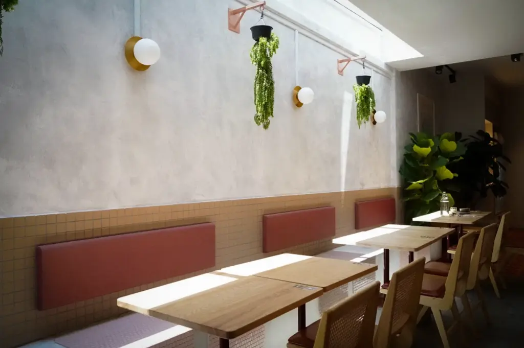 Bright minimalist dining area at Prairie by Craftsmen in Singapore featuring wooden tables, cushioned bench seating, hanging plants, and natural skylight illumination.