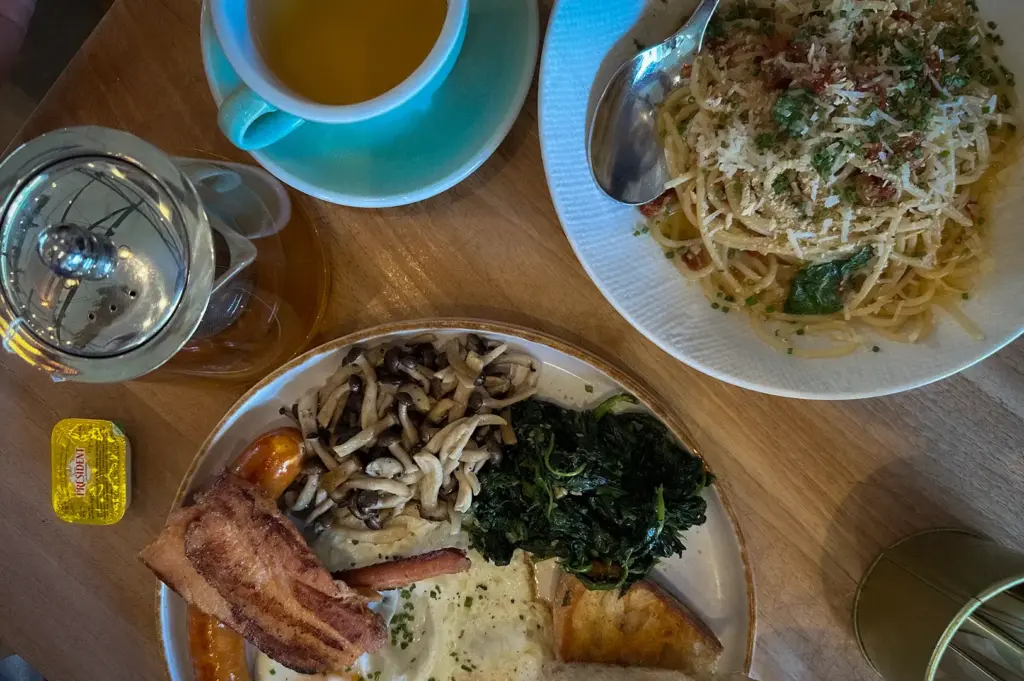 The Brewing Ground Singapore big breakfast served with eggs, sausage, bacon, mushrooms, spinach, toast, tea, and a side of pasta on a wooden table.