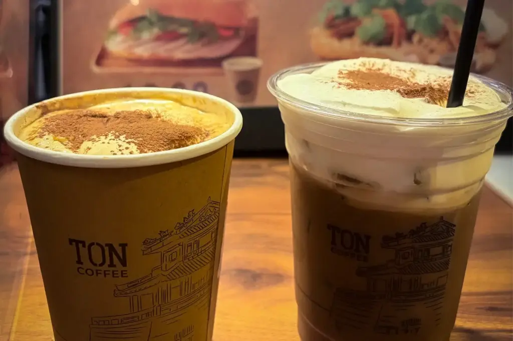 Hot and iced specialty coffee from TON Coffee Singapore, served with creamy foam and cocoa powder topping in branded takeaway cups on a wooden counter.