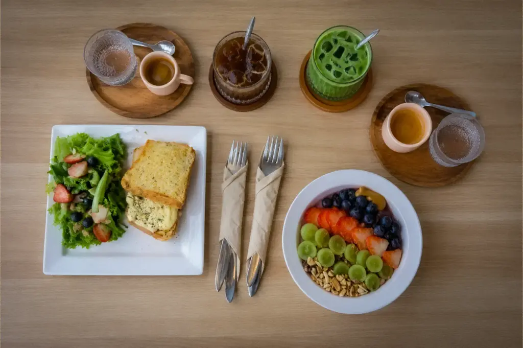 Overhead view of a brunch spread at PACTO by Parchmen Singapore featuring a sandwich with salad, acai bowl, iced matcha latte, iced coffee, and espresso served on a minimalist wooden table.
