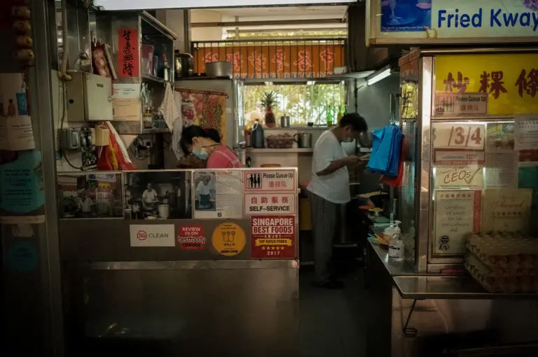 Singapore hawker stall serving Fried Kway Teow, featuring traditional signage, cooking equipment, stacked eggs, and a bustling food preparation area.