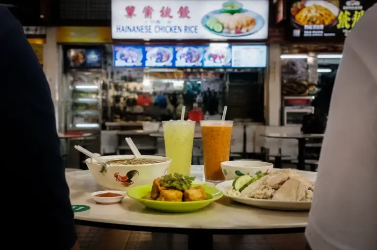 Two people dining together at a Singapore hawker centre with chicken rice, noodles, drinks, and side dishes on the table