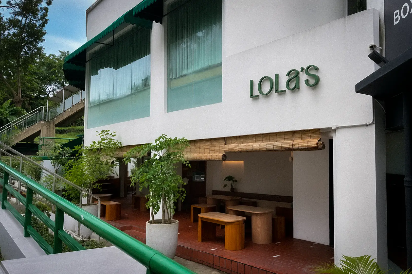 Lola’s Cafe Singapore exterior with white walls, green awnings, bamboo blinds, and outdoor seating surrounded by greenery.
