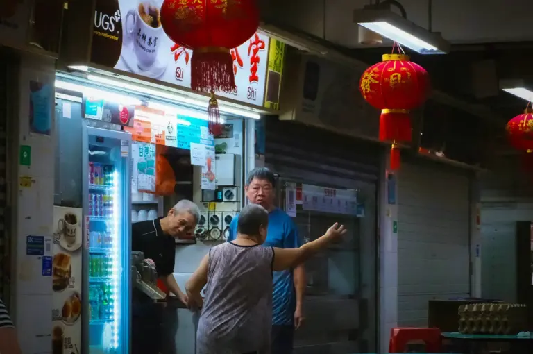 Singapore hawker stall with red Chinese lanterns, beverage counter, food preparation area, and diners inside a traditional food centre