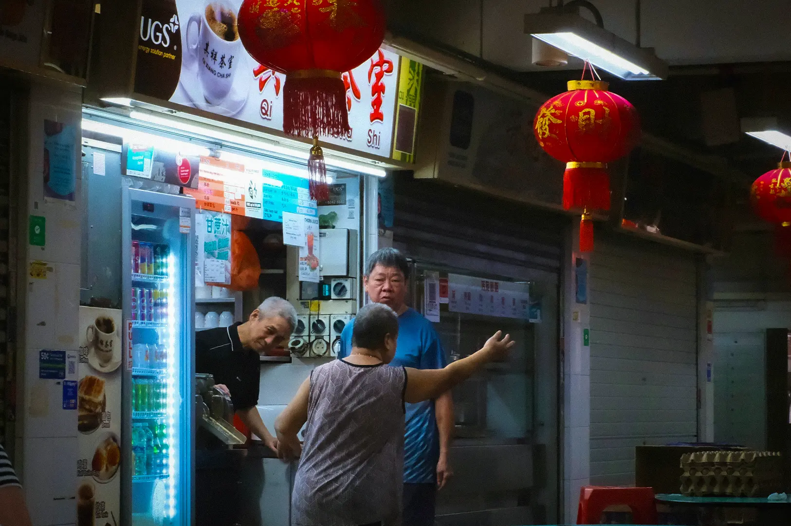 Singapore hawker stall with red Chinese lanterns, beverage counter, food preparation area, and diners inside a traditional food centre