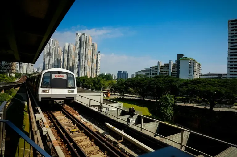 SMRT train traveling on an elevated MRT track in Singapore with modern residential towers, lush greenery, and a clear blue sky in the background.