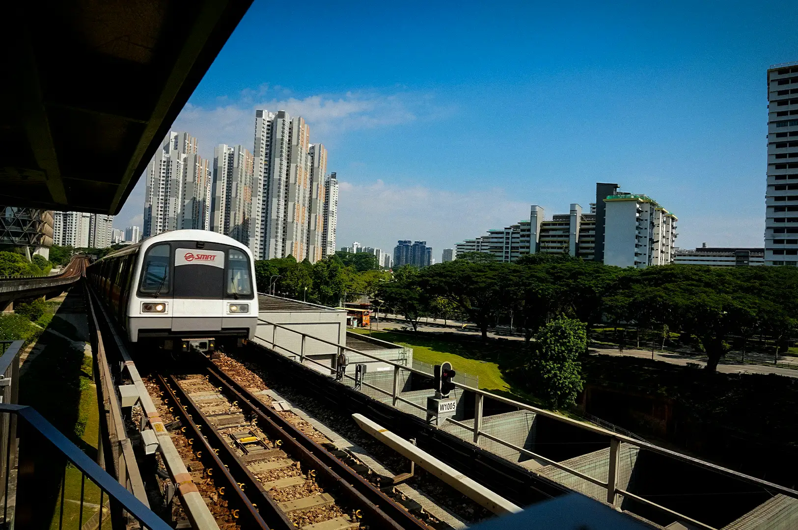 SMRT train traveling on an elevated MRT track in Singapore with modern residential towers, lush greenery, and a clear blue sky in the background.