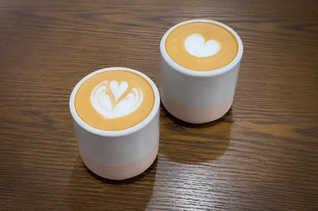 Two latte coffees with heart and floral latte art served in white ceramic cups on a wooden table