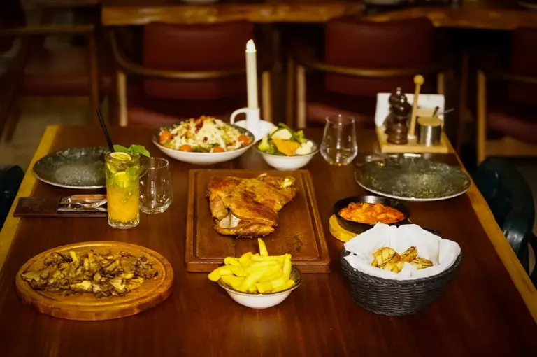 Dining table set for family dinner featuring roasted meat on a wooden board, french fries, salad, side dishes, and drinks illuminated by a candle.