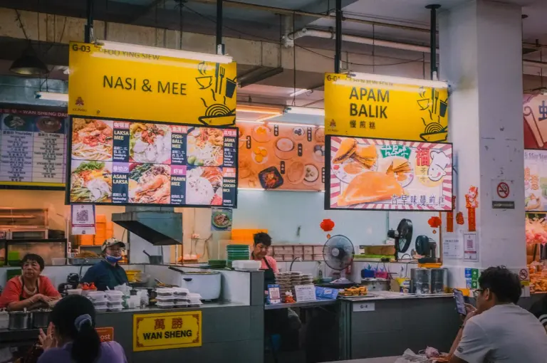 Hawker centre food stalls displaying yellow overhead menu boards offering laksa mee, fishball mee, asam laksa, prawn mee, and apam balik.