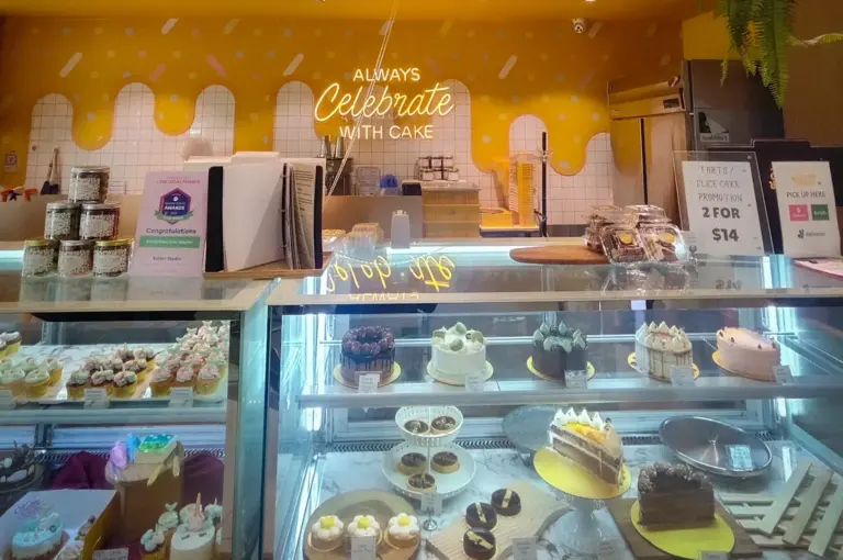 Brightly lit bakery counter displaying whole cakes, slices, and cupcakes under a neon sign reading Always Celebrate With Cake at Butter Studio in Bugis Junction, Singapore