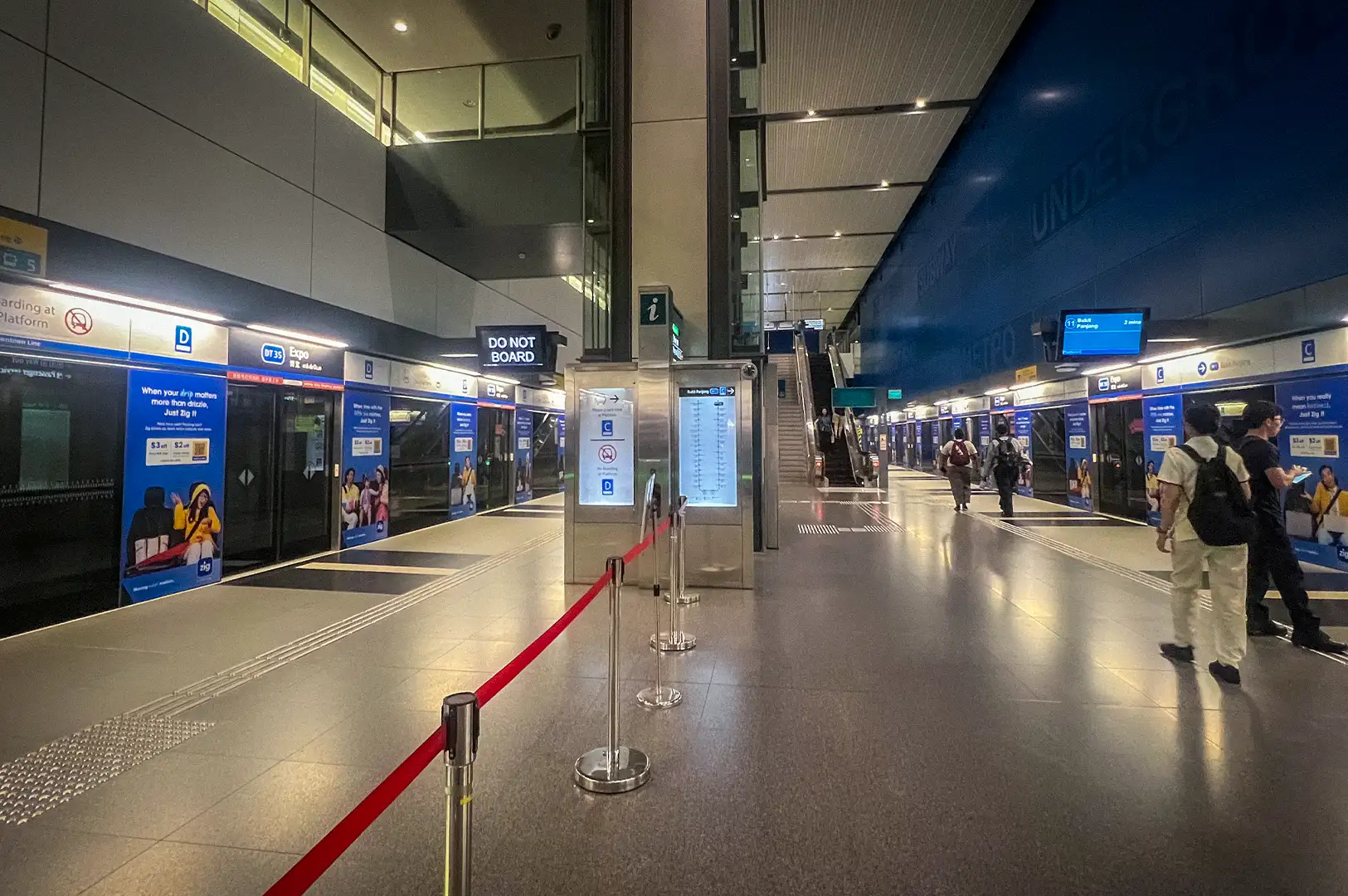 The modern platform interior of Expo MRT Station on the Downtown Line in Singapore, featuring blue-accented screen doors, clean tiled flooring, and waiting passengers.