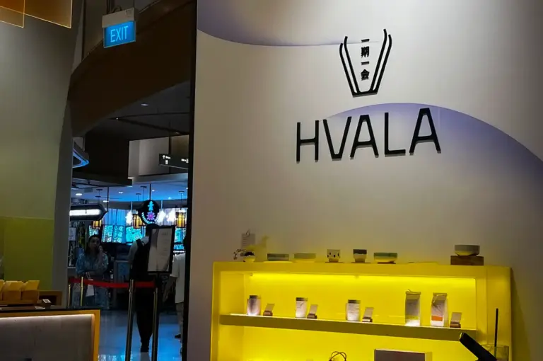 Modern interior of Hvala 111 Somerset tea house in Singapore featuring illuminated yellow display shelves with tea products and minimalist wooden tables.