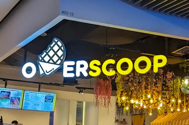 Brightly illuminated storefront sign spelling "OVERSCOOP" with a waffle-cone icon replacing the letters "V", located above a cafe interior.