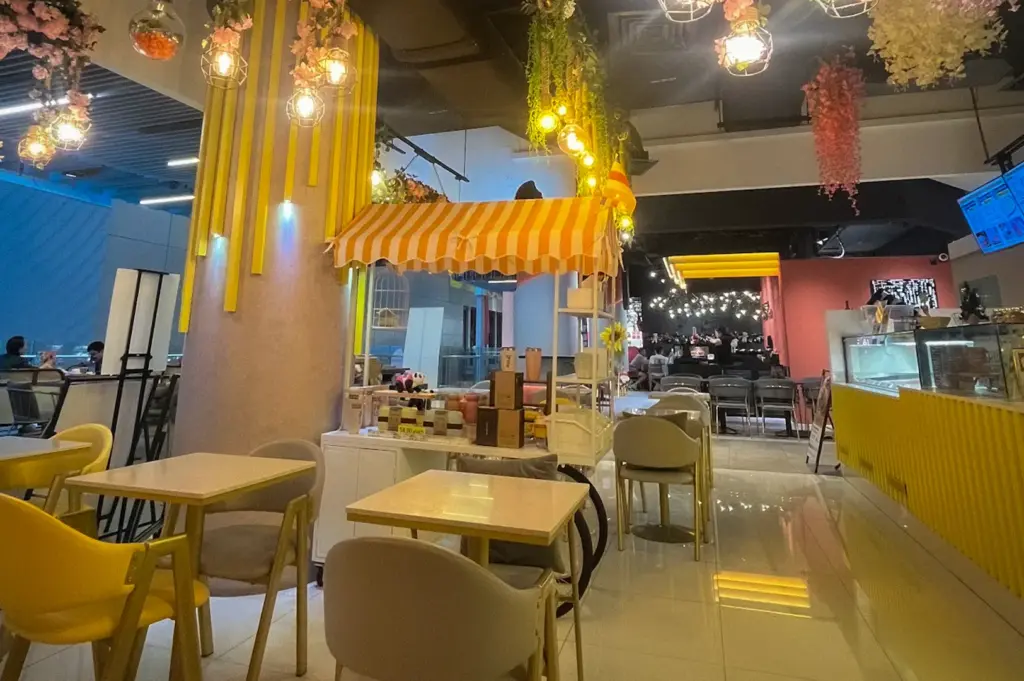 Cozy indoor dining area of Overscoop featuring hanging floral arrangements, glowing light bulbs, yellow and white tables, and modern seating.