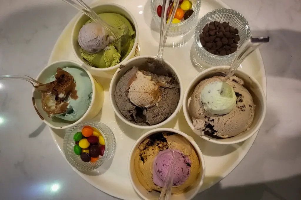 A variety flight of multiple ice cream scoops in small paper cups with clear spoons, accompanied by small glass bowls of colorful candy and chocolate chip toppings at Overscoop.
