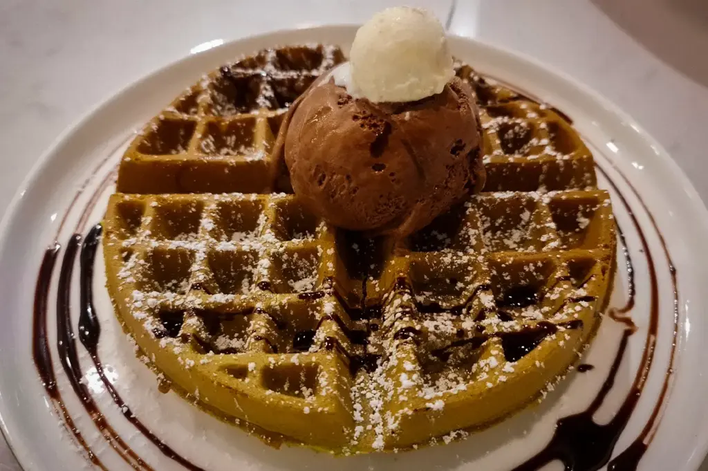 Freshly baked waffle dusted with powdered sugar and drizzled with chocolate sauce, topped with a scoop of chocolate ice cream and vanilla cream at Overscoop.