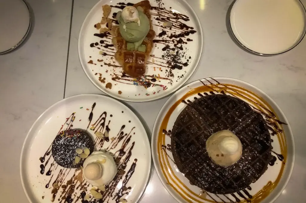 A top-down view of a spread of desserts at Overscoop featuring chocolate waffles, a lava cake with ice cream, and a croiffle topped with green tea ice cream, all drizzled with chocolate and caramel sauce.