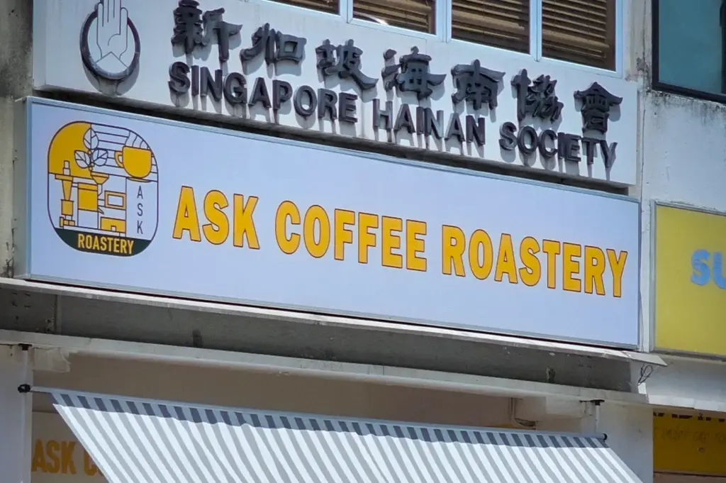 ASK Coffee Roastery Singapore storefront with striped awning, outdoor cafe seating, potted plants, and Singapore Hainan Society signage above the entrance