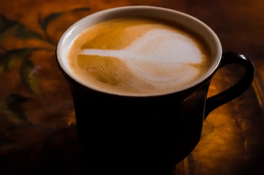 A close-up shot of a dark ceramic cup filled with a warm latte featuring delicate white foam art, resting on a textured wooden surface.