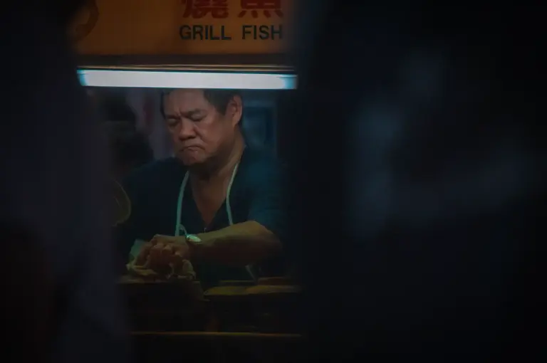 A focused local hawker vendor cooking food at a grill fish and seafood stall illuminated under bright overhead stall lighting