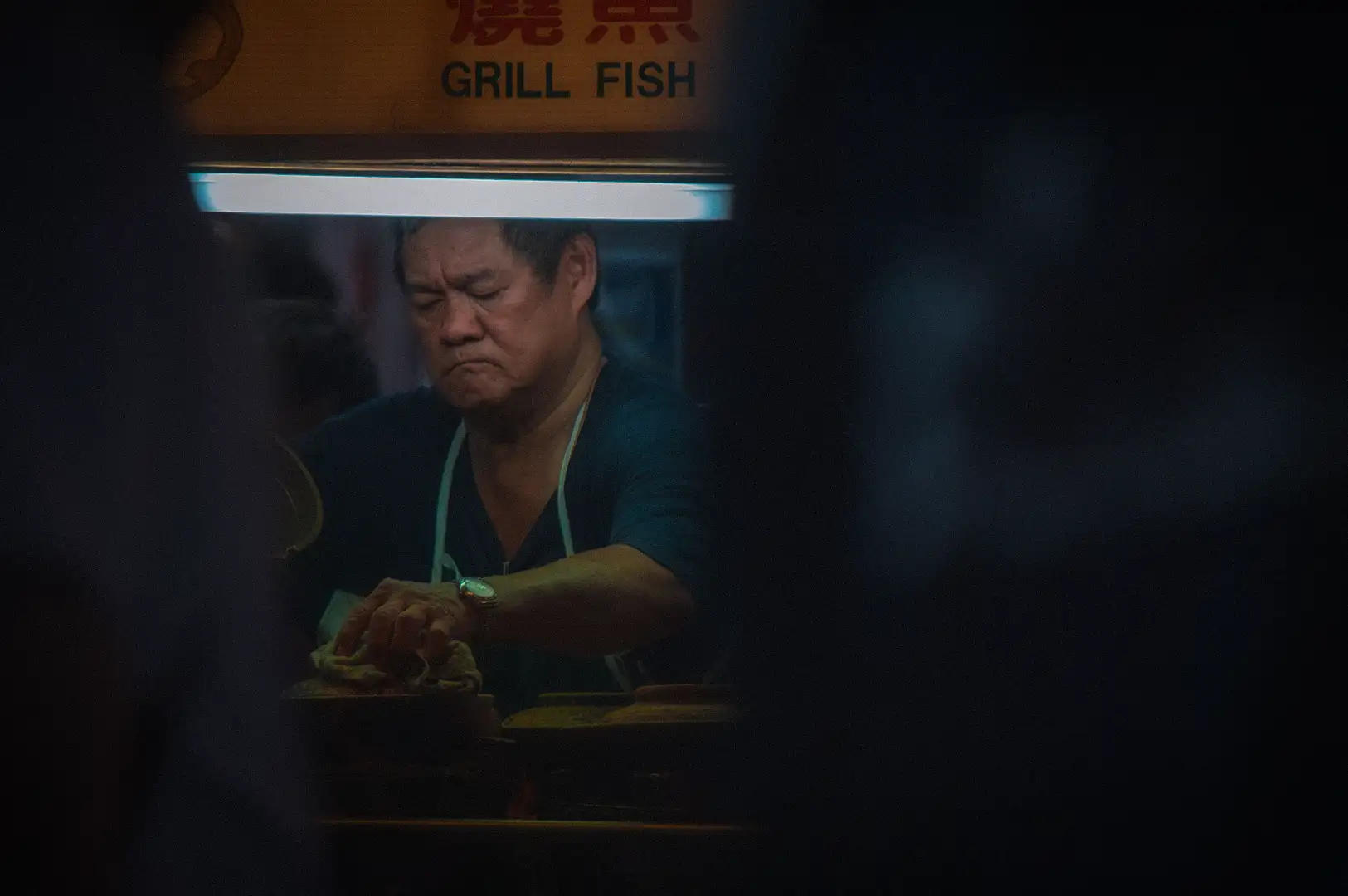 A focused local hawker vendor cooking food at a grill fish and seafood stall illuminated under bright overhead stall lighting