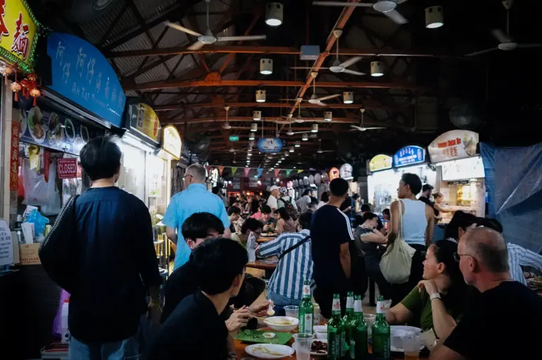 Bustling hawker centre food market interior in Singapore featuring rows of food stalls and a lively crowd dining at tables beneath a high wooden ceiling