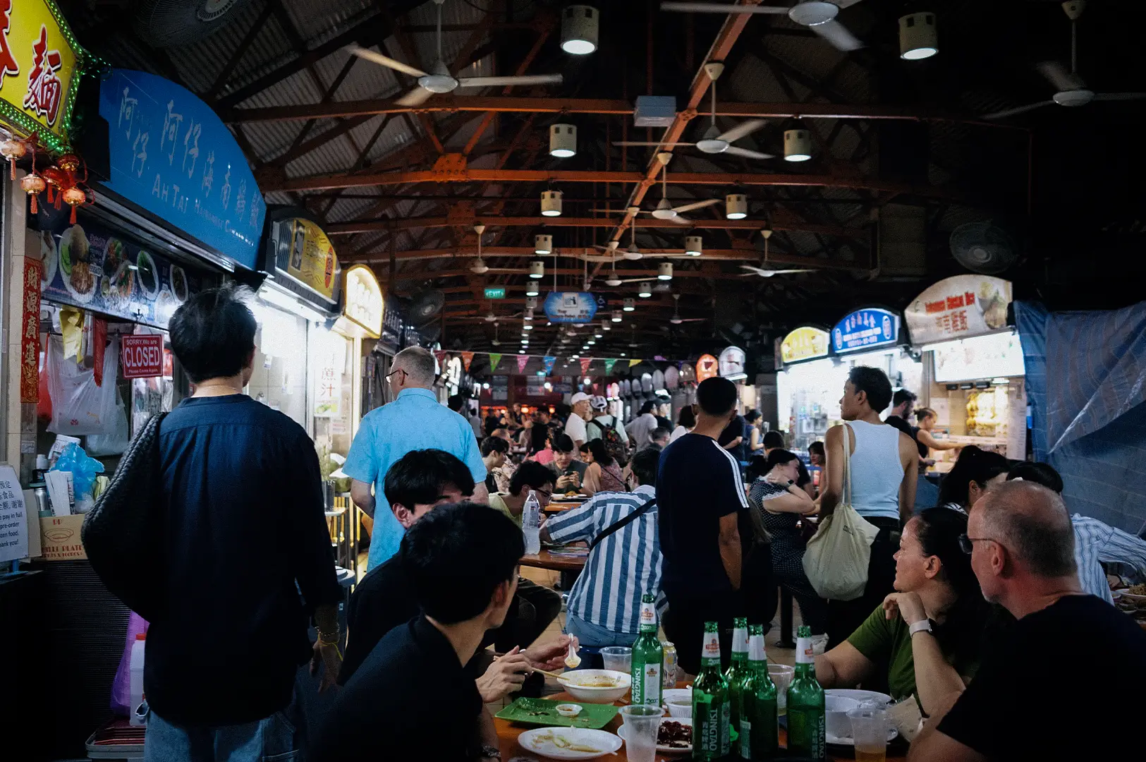 Bustling hawker centre food market interior in Singapore featuring rows of food stalls and a lively crowd dining at tables beneath a high wooden ceiling