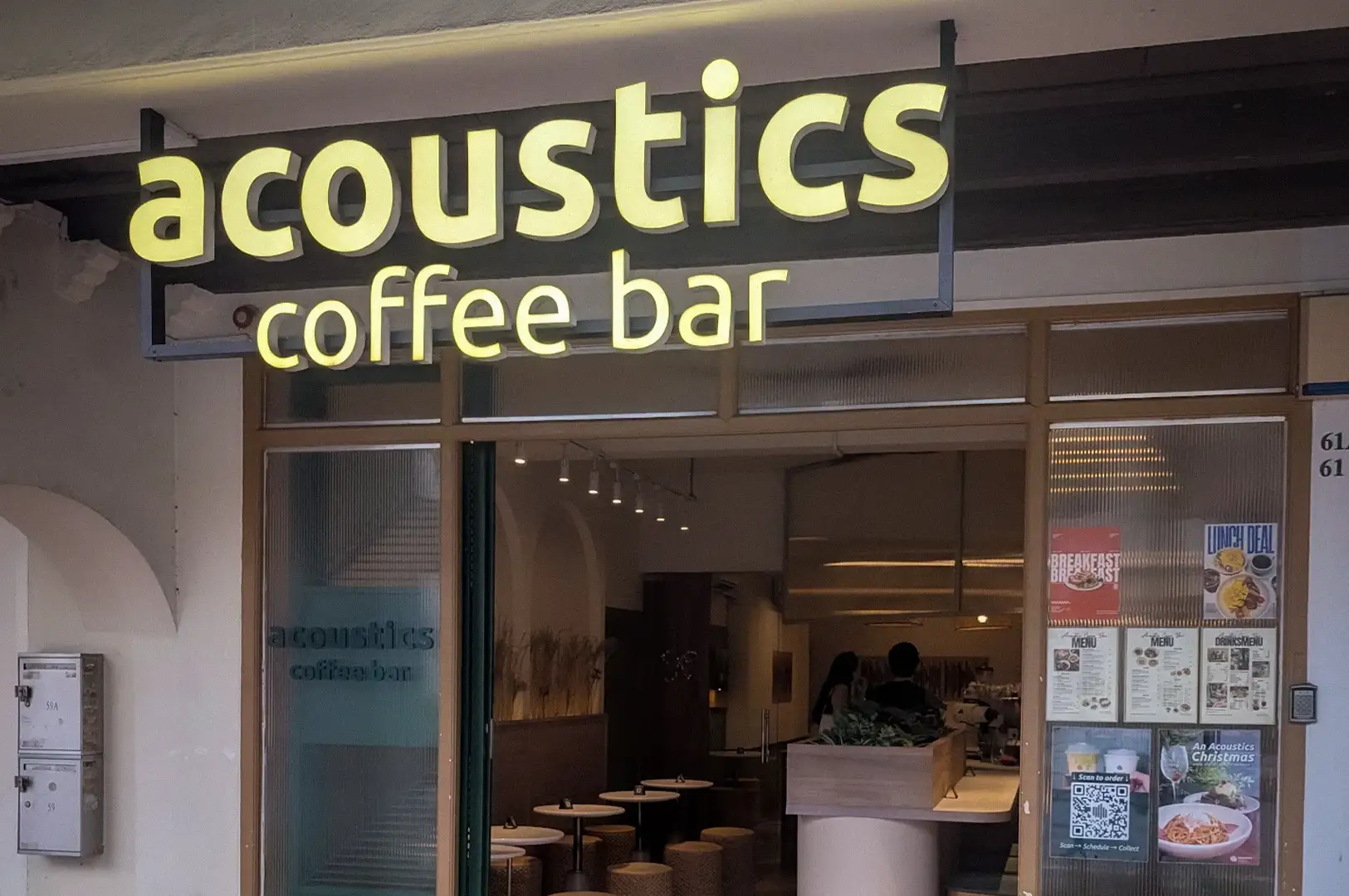Illuminated storefront sign and open entrance of Acoustics Coffee Bar along Neil Road in Singapore, featuring rattan benches and a cozy interior view