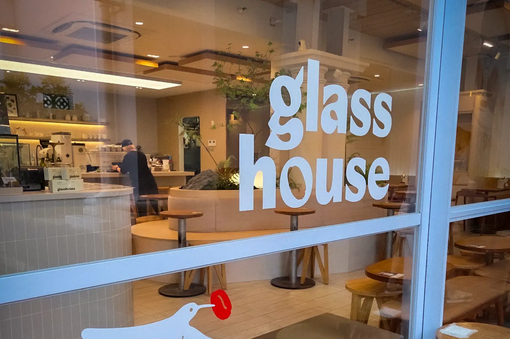 Exterior glass window view into the bright, minimalist interior of Glasshouse cafe at CHIJMES with wooden seating and bird decal in Singapore