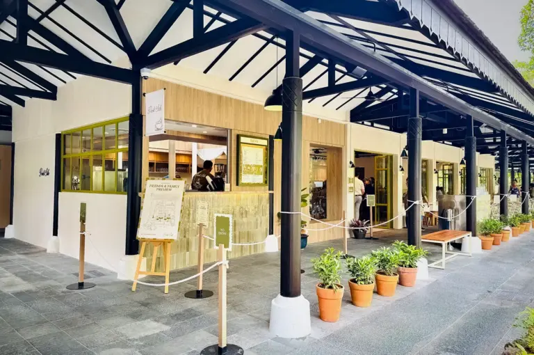 Exterior storefront and sheltered walkway of Park Side Cafe featuring a covered veranda, wooden pillars, potted plants in terracotta pots, and queue stanchions