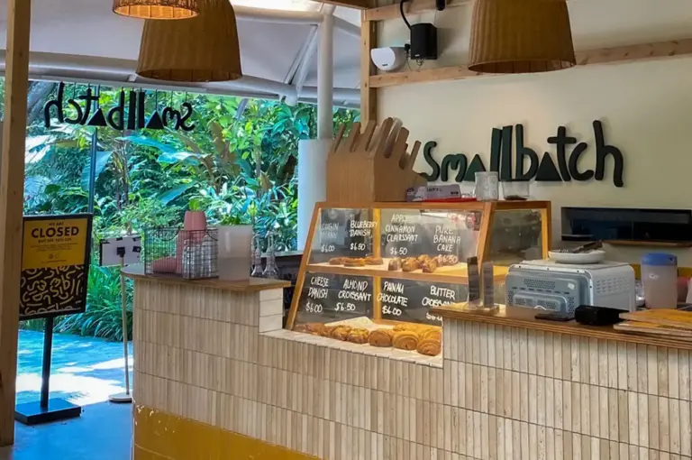 An open-air counter view of Small Batch cafe at the Singapore Botanic Gardens, featuring woven pendant lamps, a tiled ordering counter with pastry display cases, and lush greenery outside.
