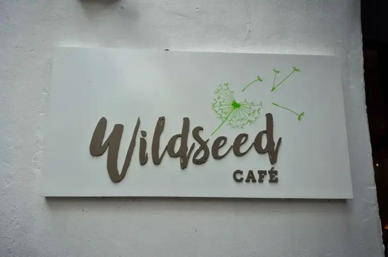 Wildseed Café signage featuring dark lettering and a green dandelion logo mounted on a textured white wall