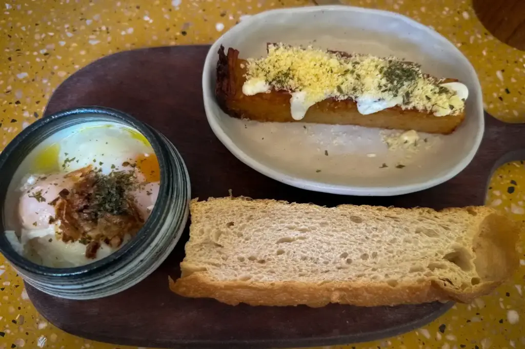An overhead view of a breakfast dish at Small Batch cafe in Singapore, featuring soft-boiled eggs topped with crispy bits in a small bowl, crusty artisanal bread, and a side of seasoned potato cake served on a wooden board.