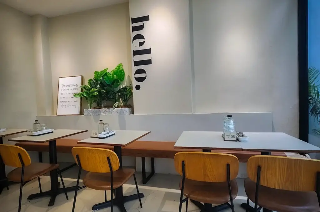 Minimalist dining interior at Daily Ground in Bukit Timah, featuring wooden dining chairs, white tables, leather banquette seating, and a feature wall with "hello." lettering