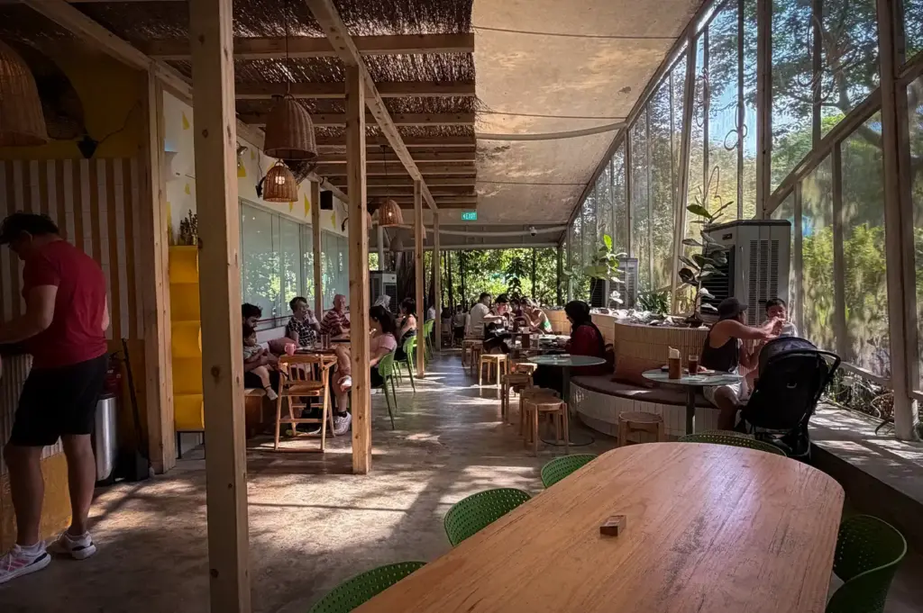 An indoor dining view of Small Batch cafe in Singapore, featuring wooden communal tables, floor-to-ceiling glass windows overlooking greenery, and patrons seated throughout the space.