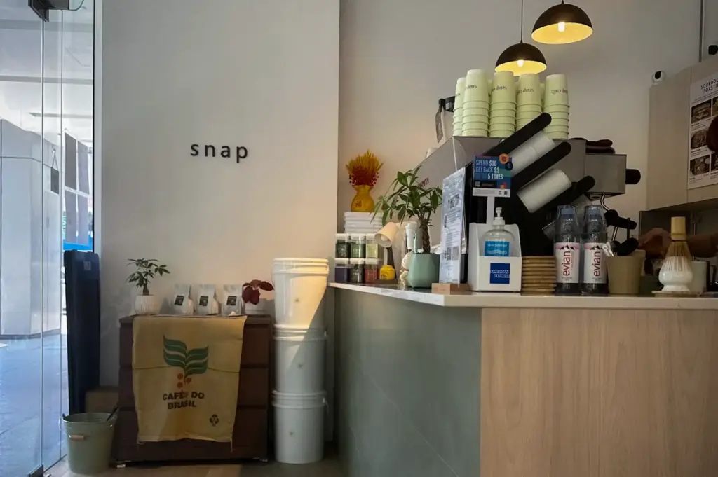 Interior view of Snap Cafe (The Clift) - Specialty Coffee, Freshly-whisked Matcha, Sourdough Toasties highlighting the minimalist coffee counter, pendant lighting, stacked paper cups, and a branded "snap" wall sign