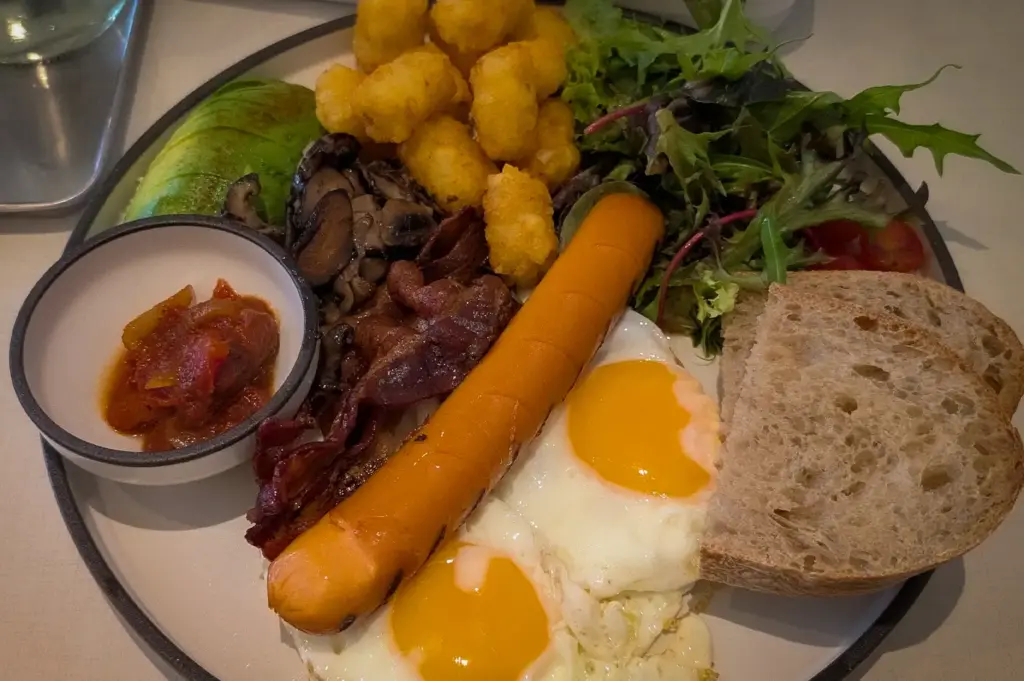 A hearty big breakfast platter served at Daily Ground, featuring sunny-side-up eggs, a grilled sausage, crispy tater tots, sliced sourdough bread, avocado, mushrooms, bacon, and side salad