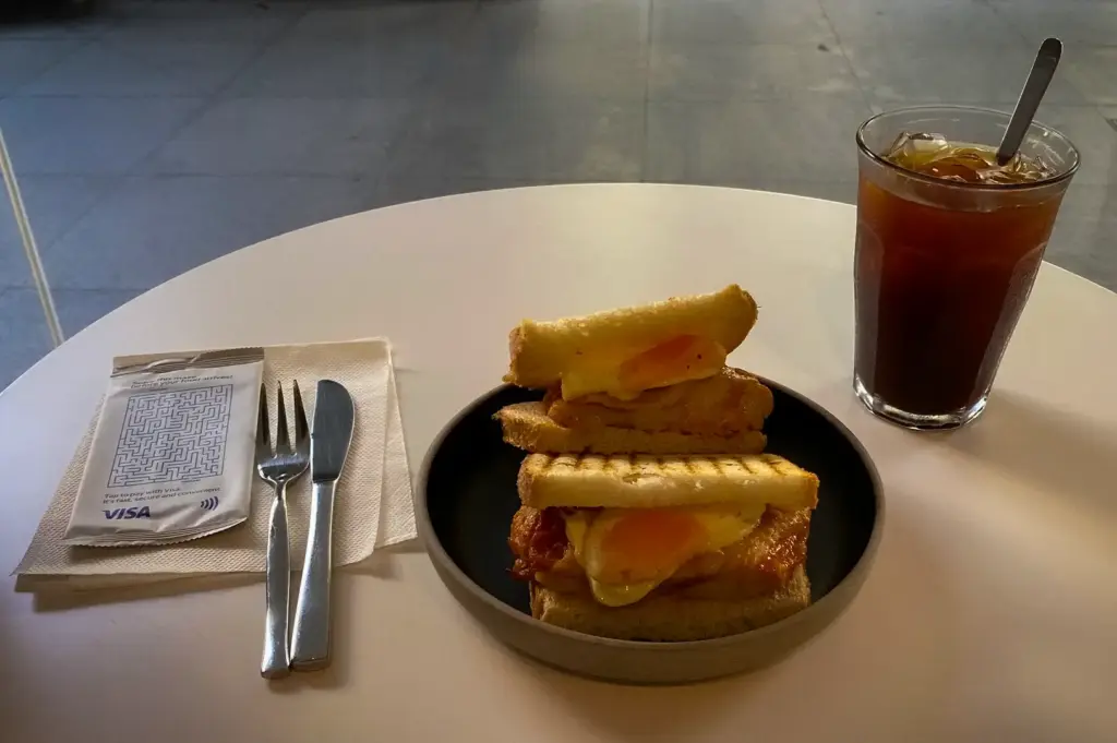 A grilled chicken and egg sourdough toastie cut in half served on a dark plate alongside a glass of iced long black coffee on a cafe table at Snap Cafe (The Clift) - Specialty Coffee, Freshly-whisked Matcha, Sourdough Toasties