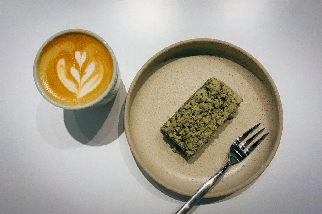 A slice of matcha crumb cake on a ceramic plate with a small fork alongside a hot latte with latte art served on a white table at Snap Cafe (The Clift) - Specialty Coffee, Freshly-whisked Matcha, Sourdough Toasties