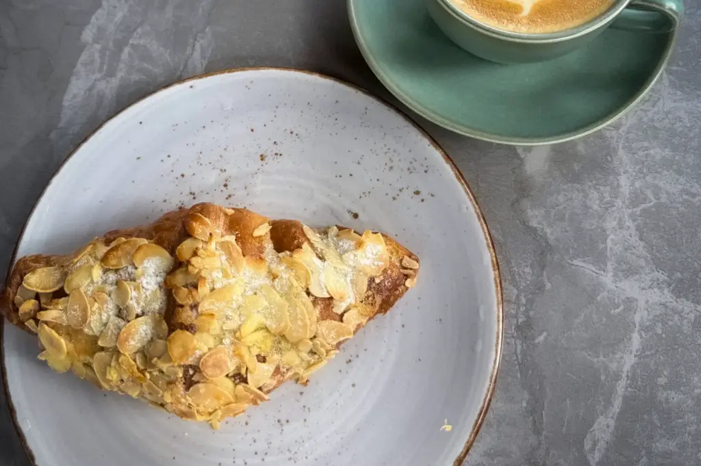 Wildseed Café serving featuring a flaky almond croissant on a plate alongside a warm latte in a green cup with heart latte art on a marble table