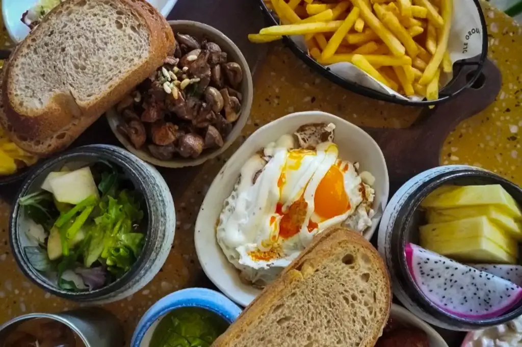 An overhead view of a hearty breakfast platter at Small Batch cafe in Singapore, featuring artisanal bread, fried eggs, sausages, sautéed mushrooms, French fries, fresh fruit, and iced coffee arranged on a yellow-patterned table.