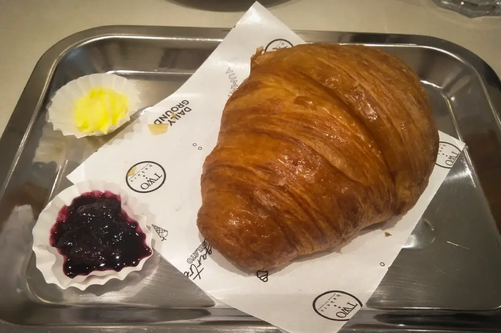 A golden-brown baked butter croissant served on a metal tray with butter and berry jam at Daily Ground